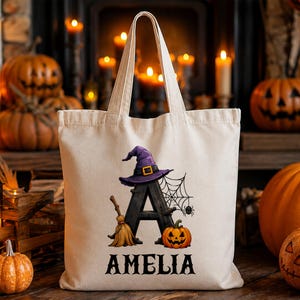 May include: A beige canvas tote bag featuring a large black letter A decorated with a purple witch hat, a broomstick, a spider web, and a small orange jack-o'-lantern. The name AMELIA is printed in black capital letters below the graphic.