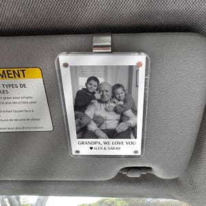 May include: A clear acrylic photo frame with a black and white image of a grandfather and two grandchildren. The text "GRANDPA, WE LOVE YOU" and "ALEX & SARAH" is printed at the bottom. The frame is attached to a car visor.