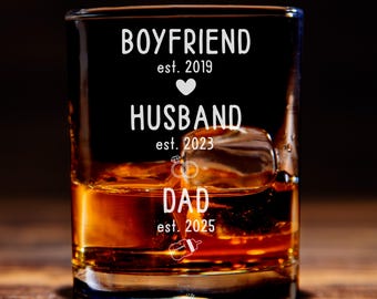 May include: A clear glass with a brown liquid inside. The glass has the text "Boyfriend est. 2019" at the top, "Husband est. 2023" in the middle, and "Dad est. 2025" at the bottom.