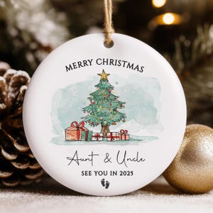 May include: White round Christmas ornament with a watercolor illustration of a Christmas tree with presents underneath. The text reads "Merry Christmas Aunt & Uncle See You in 2025" with a footprint symbol.