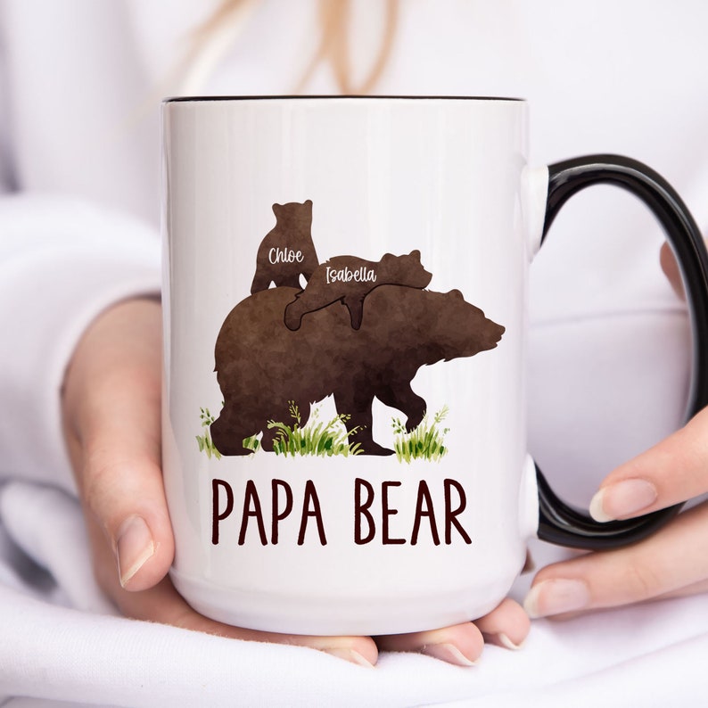 K&ouml;nnte beinhalten: Eine wei&szlig;e Keramiktasse mit schwarzem Henkel. Die Tasse zeigt eine braune Aquarell-Illustration eines B&auml;ren mit zwei Jungen auf seinem R&uuml;cken. Die Jungen sind mit "Chloe" und "Isabella" beschriftet. Der Text "Papa Bear" ist unter dem B&auml;ren gedruckt.