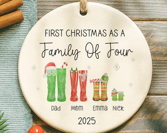 May include: Round ceramic Christmas ornament with a family-themed design. The ornament features green and red boots representing family members, with the text "First Christmas As A Family Of Four" and the year 2025.