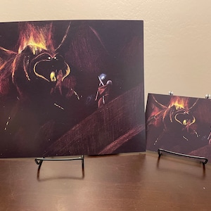 May include: A black and white drawing of a fiery dragon with two figures in the foreground. The dragon is depicted with a fiery mane and wings, and its eyes glow yellow. The figures are small in comparison to the dragon and appear to be facing it.