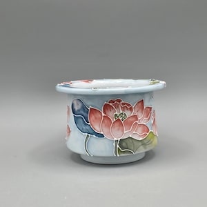 May include: A light blue ceramic bowl featuring a hand-painted design of pink lotus flowers and green leaves. The bowl has a wide, flat rim and a tapered base, with white outlines defining the floral motifs against the pale blue background.