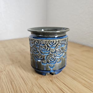 May include: A ceramic planter with a dark gray exterior and a blue interior. The planter features a repeating rose pattern carved into the surface. The rim and base are also blue, creating a cohesive design.