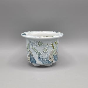 May include: A small, round, light blue ceramic planter with a decorative floral design in shades of blue, green, and tan. The planter has a scalloped rim and sits on three small feet.
