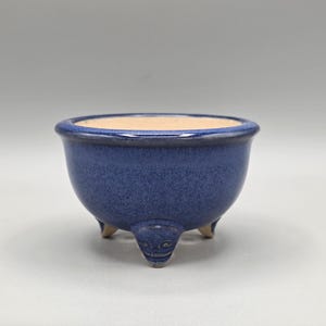 May include: A small, round ceramic planter with a deep blue, speckled glaze finish. The bowl features a smooth, rounded rim and is supported by three short, sculpted feet that have a subtle, carved facial detail on the front-facing leg.