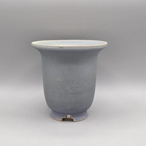 May include: A light blue ceramic pot with a speckled finish and a flared rim. The vessel features a rounded body that tapers toward a small, notched base. The surface has a matte texture with subtle variations in color and a light-colored rim edge.