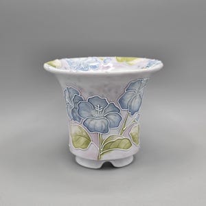 May include: A ceramic vase with a flared rim and a footed base. The vase is decorated with blue floral designs and green leaves against a light purple background. The rim has a textured, light blue and green pattern.