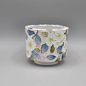 May include: A cylindrical ceramic cup featuring a raised, textured floral pattern with white blossoms and blue-tinted leaves against a soft pink background. The rim has an uneven, organic edge, and the interior is a smooth, glossy white finish.