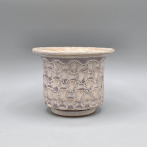 May include: A ceramic planter with a woven pattern design. The planter is a light purple color with a beige rim and base. The planter has a round shape and is suitable for indoor or outdoor use.