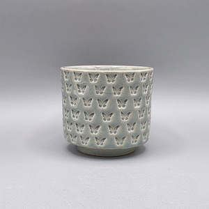 May include: A light blue ceramic pot with a repeating butterfly pattern. The pot has a cylindrical shape with a slightly raised base. The butterflies are embossed, creating a textured surface.