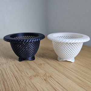 May include: Two small, decorative mesh bowls. One is black, the other is white. Both have a woven rim and a flared, perforated body. The bowls sit on a wooden surface.