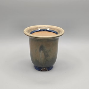 May include: A ceramic vase with a flared rim and a rounded base. The vase has a gradient of colors, transitioning from a light tan at the rim to a darker blue-green at the base. A thin blue band accents the rim.