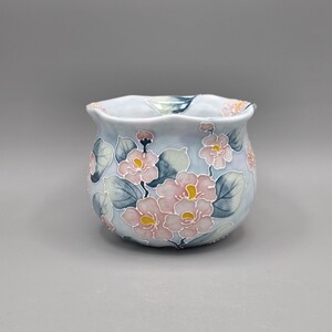 May include: A small, rounded ceramic bowl with a light blue base color, featuring a hand-painted floral pattern of soft pink blossoms with yellow centers and muted green leaves, finished with a scalloped, wavy rim edge.