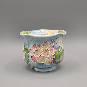 May include: A small ceramic bowl with a light blue background featuring hand-painted pink and white lotus flowers and green leaves. The bowl has a scalloped rim and a rounded body, showcasing detailed floral motifs with raised outlines.