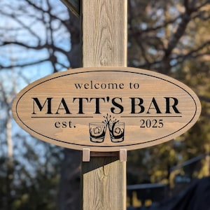 May include: Wooden oval sign with the text "welcome to MATT'S BAR est. 2025" in black lettering. The sign features two glasses with ice cubes and is mounted on a wooden post.
