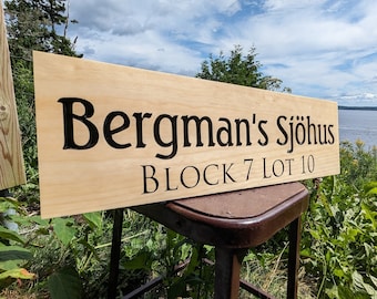 May include: A rectangular wooden sign with the text "Bergman's Sjöhus BLOCK 7 LOT 10" in black lettering. The sign is made of light-colored wood and is displayed outdoors, with a scenic background of water and sky.