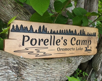 Puede incluir: Un letrero de madera con letras negras que dice "Porelle's Camp - Oromocto Lake". El letrero tiene una silueta de un bosque en la parte superior y un colimbo nadando en un lago en la parte inferior.