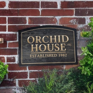 Pu&ograve; includere: Un cartello in legno nero con un'iscrizione dorata che recita "Orchid House Established 1982". Il cartello &egrave; montato su un muro di mattoni.