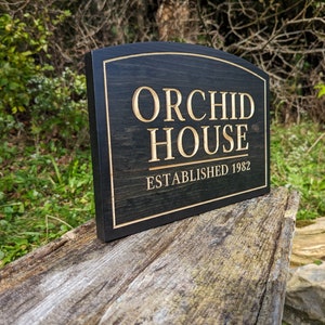 Pu&ograve; includere: Un cartello in legno nero con bordi arrotondati e lettere dorate. Il cartello recita "Orchid House Established 1982".