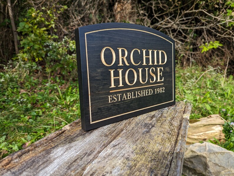 Pu&ograve; includere: Un cartello in legno nero con bordi arrotondati e lettere dorate. Il cartello recita "Orchid House Established 1982".