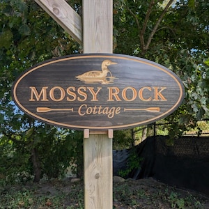 May include: Dark brown wooden sign for "Mossy Rock Cottage" with a loon illustration and gold lettering. The oval sign has a gold border and is mounted on a wooden post.