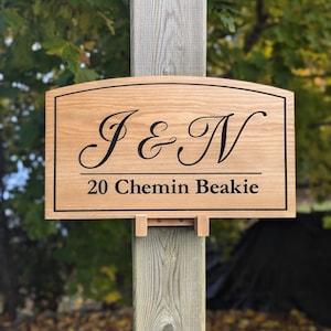 Pu&ograve; includere: Un cartello in legno con la parte superiore curva, che presenta le iniziali "J & N" in un carattere corsivo e l'indirizzo "20 Chemin Beakie" sotto. Il cartello &egrave; montato su un palo di legno.