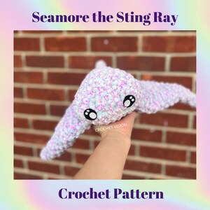 May include: A crocheted stingray plush toy in shades of purple, pink, and white. The toy has large black eyes and is held in a hand against a brick wall. The text "Seamore the Sting Ray Crochet Pattern" is visible below the toy.