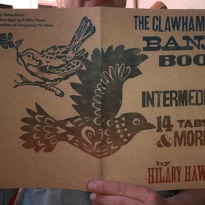 以下が含まれることがあります： クローハンマー・バンジョー・ブック：中級、14のタブ譜とその他、ヒラリー・ホーク著。表紙は茶色の背景に、花が咲いた枝にとまっている黒い鳥と、飛んでいる黒い鳥の2羽が描かれている。タイトルは青と赤の文字で書かれている。