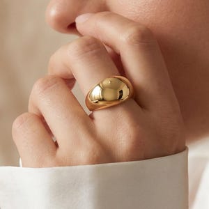 May include: A polished gold ring with a smooth, rounded, and domed surface. The ring features a thick, solid band design that tapers slightly toward the palm, showcasing a high-shine metallic finish that reflects light across its curved exterior.