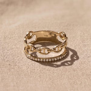 May include: A gold ring featuring a dual-band design with a top chain-link section and a bottom band encrusted with small clear stones. The interior of the ring is clearly stamped with the text 14k, indicating the gold purity of the metal.