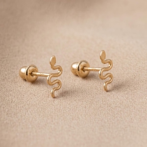 Könnte beinhalten: Ein Paar goldene Ohrstecker mit einem schlangenförmigen Design. Jeder Ohrstecker verfügt über einen Stift mit Schraubverschluss. Das Metall hat eine polierte Oberfläche und das Schlangenmotiv ist als einfache, durchgehende, gewundene Linie dargestellt.