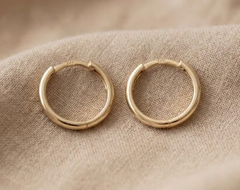 May include: A pair of small, polished gold hoop earrings with a smooth, circular design. Each earring features a subtle hinge mechanism for closure and a tiny 14k stamp visible on the metal surface, reflecting a bright, warm metallic yellow tone.