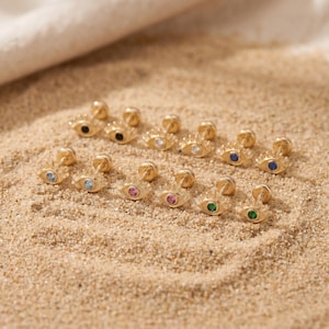May include: A collection of gold-toned stud earrings shaped like eyes, each featuring a small central gemstone. The stones vary in colour, including black, clear, blue, pink, and green, with radiating metal lines forming the eye shape.