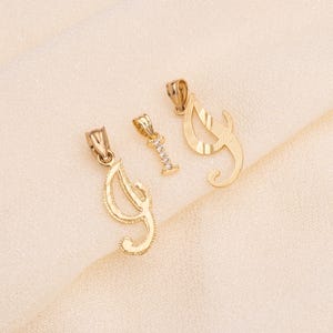 Puede incluir: Tres colgantes dorados sobre una tela color crema. Dos colgantes son formas estilizadas de la letra "J", y el tercero es una "I" con pequeñas piedras transparentes. Los colgantes son probablemente joyas, posiblemente para collares o pulseras.