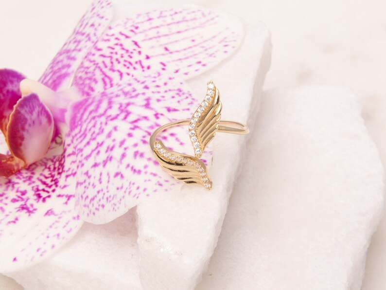 May include: A gold ring with a design of two wings with clear stones. The ring is on a white surface with a pink flower.
