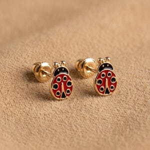 May include: A pair of 14k gold stud earrings shaped like ladybirds. Each earring features a red enamel body with black spots and a black head. The earrings include gold screw-back posts, with the 14k hallmark clearly visible on the metal backing.