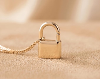 May include: A gold-toned metal padlock pendant attached to a delicate box chain necklace. The padlock features a smooth, polished surface with a rounded shackle and a rectangular body, reflecting light across its metallic finish.