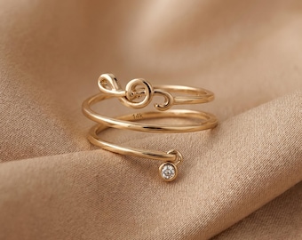 Può includere: Un anello in oro con un design a chiave di violino su un'estremità e una piccola pietra preziosa rotonda e trasparente sull'altra. La fascia ha una forma a spirale con l'incisione 14k visibile sulla superficie interna. Il metallo è lucido.