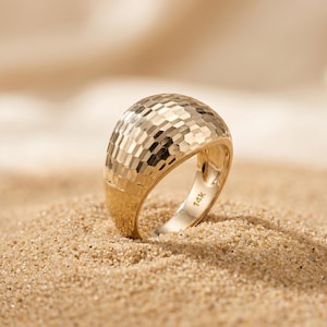 Könnte beinhalten: Ein breiter, gewölbter Goldring mit einer facettierten, wabenartigen Textur auf der Oberfläche. Die Innenseite des Rings ist deutlich mit dem Stempel 14K versehen, was den Goldgehalt dieses polierten und reflektierenden Schmuckstücks angibt.
