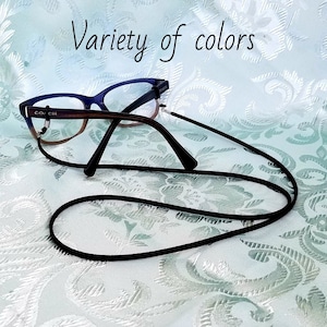 May include: Black eyeglass chain with a pair of glasses on a white patterned surface. The glasses have a brown frame and blue lenses. The text "COACH" is visible on the frame.