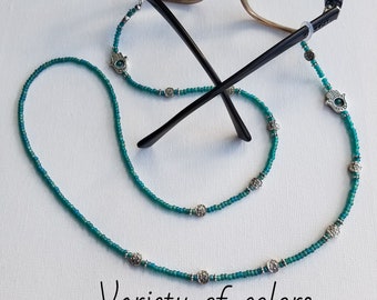May include: Teal blue beaded eyeglass chain with silver hamsa hand charms. The chain is attached to the arms of a pair of eyeglasses.