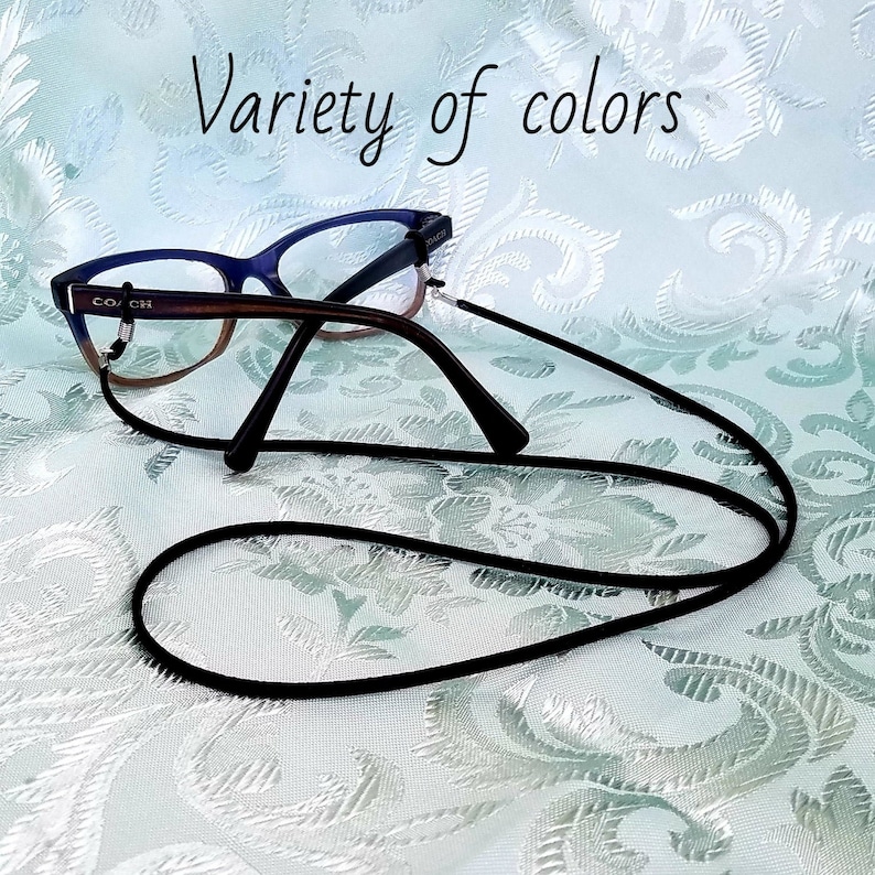 May include: Black eyeglass chain with a pair of glasses on a white patterned surface. The glasses have a brown frame and blue lenses. The text "COACH" is visible on the frame.
