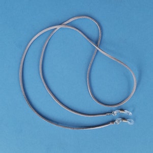 May include: A pair of gray suede eyeglass holders with silver metal clasps. The holders are looped together and laid out on a blue background.