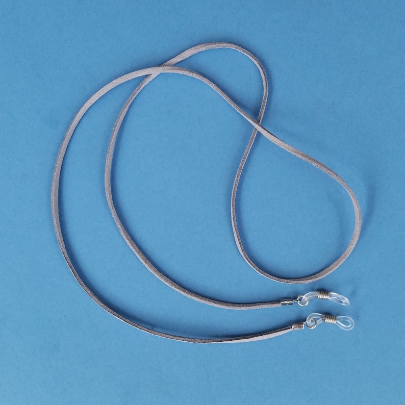 May include: A pair of gray suede eyeglass holders with silver metal clasps. The holders are looped together and laid out on a blue background.
