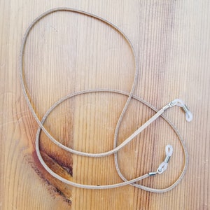 May include: A light brown leather eyeglass chain with silver metal clasps. The chain is lying on a wooden surface.