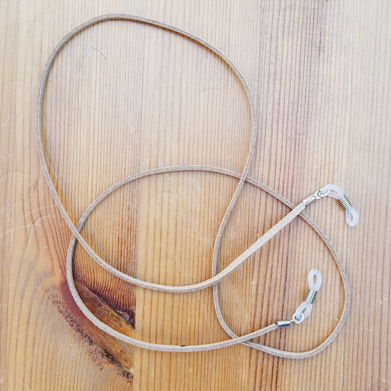 May include: A light brown leather eyeglass chain with silver metal clasps. The chain is lying on a wooden surface.