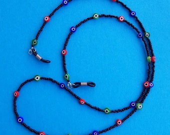 May include: A beaded eyeglass chain featuring small black seed beads interspersed with round, multi-colored evil eye beads in red, blue, and green. The chain includes black rubber loops at both ends with silver-toned metal coils for attachment.