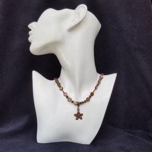 May include: A brown and white beaded necklace with a copper flower pendant. The necklace is on a white mannequin.